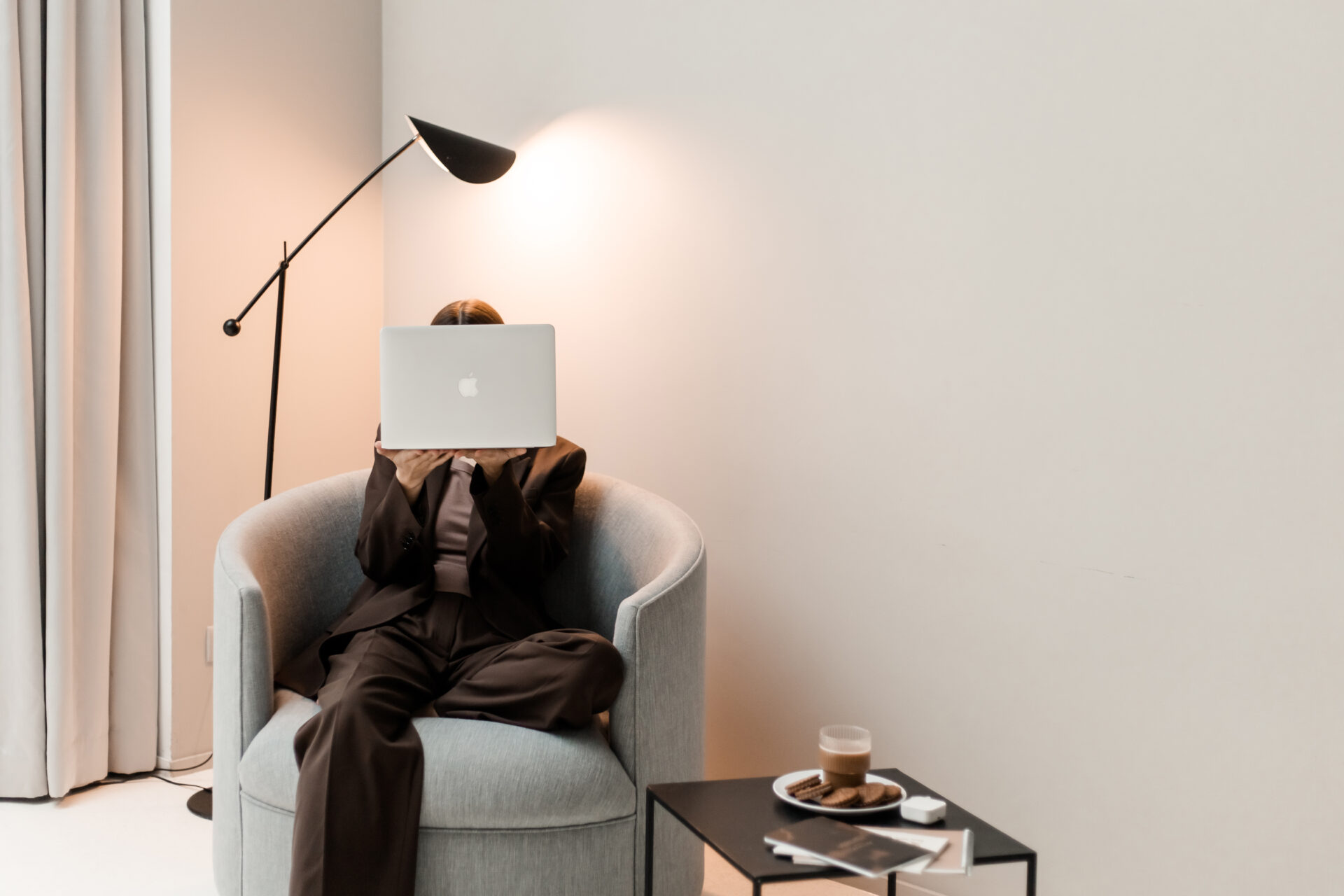 Frau in braunem Outfit sitzt mit Laptop in einem hellen Sessel. Minimalistischer Arbeitsplatz mit Stehlampe, Kaffee und Beistelltisch.