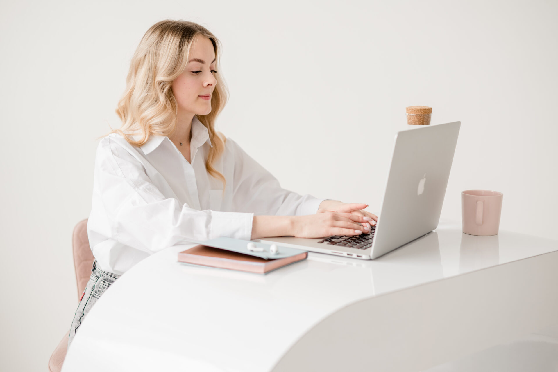 Frau sitzt an einem aufgeräumten Schreibtisch und arbeitet am Laptop. Helles, minimalistisches Setting mit Notizbuch und Kaffeetasse.