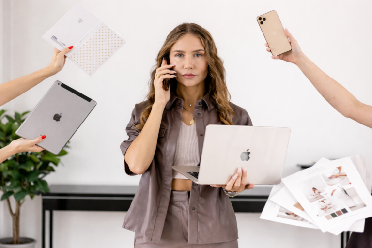 Soloselbständige steht mit Laptop, Smartphone und Unterlagen und wird von mehreren Aufgaben gleichzeitig beansprucht.